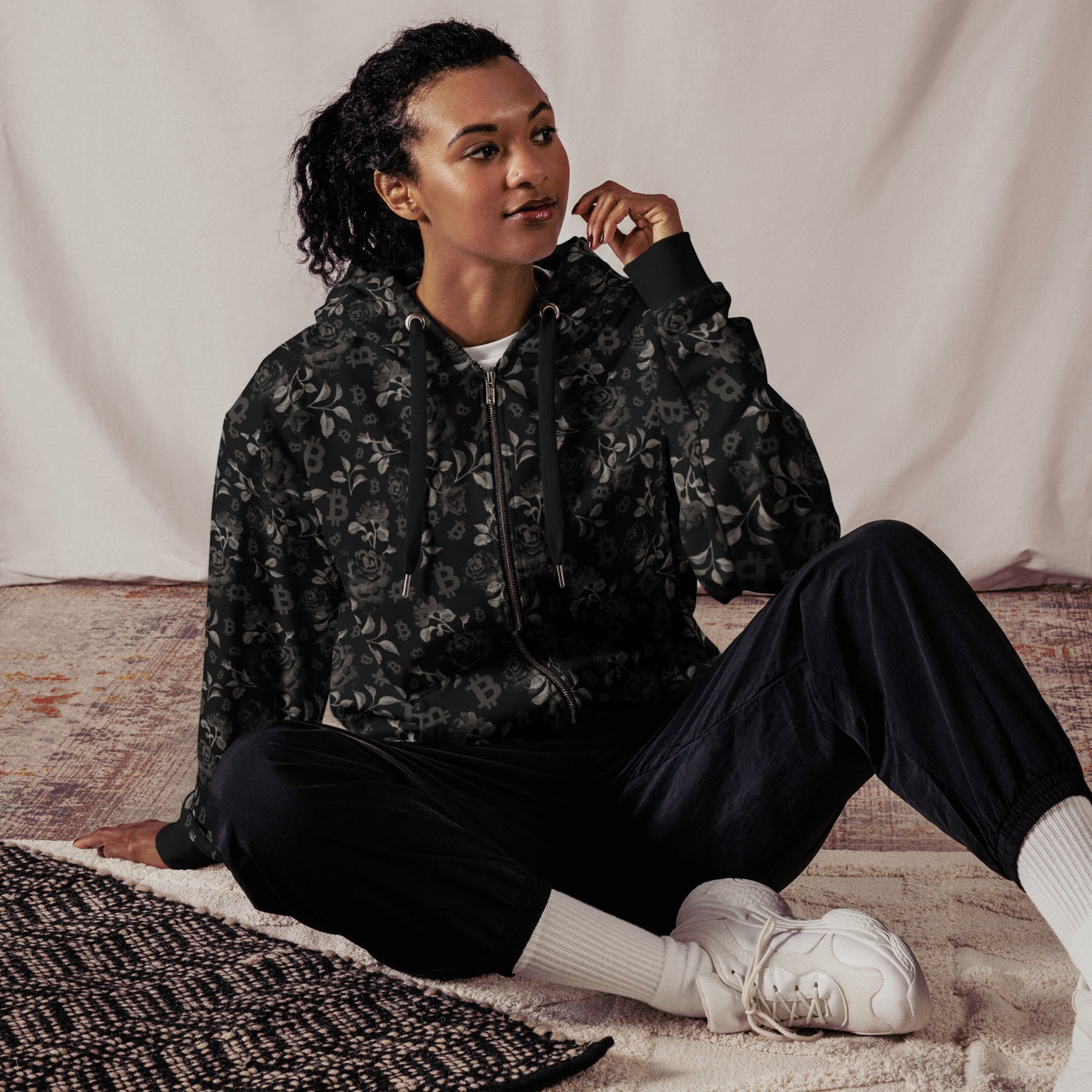 Person wearing a black floral bitcoin hoodie and pants sitting on a textured rug.